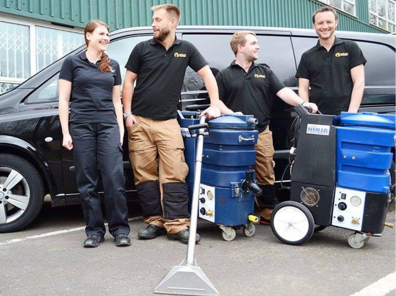 This is a photo of Clapham Carpet Cleaning Solutions carpet cleaners (three men and one woman) standing in fromt of their black van, with two steam cleaning carpet machines next to them